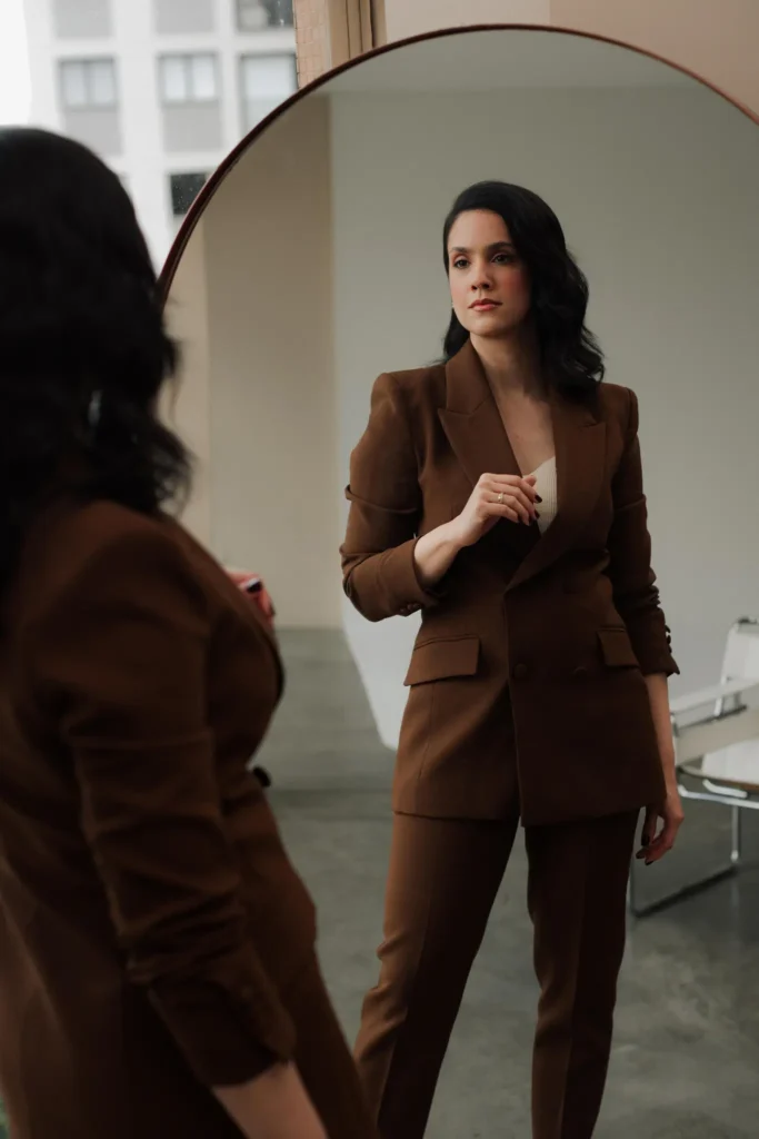 Cirurgiã plástica Roberta Araújo se olhando em um espelho e usando um conjunto de blazer e calça de alfaiataria marrom